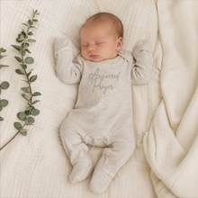 Load image into Gallery viewer, A sleeping newborn baby wearing a light heather gray footed bodysuit with 'Answered Prayer' printed in gray script on the chest, lying on a textured cream blanket next to a green eucalyptus branch.
