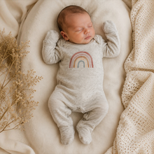 Load image into Gallery viewer, A sleeping newborn baby wearing a light heather oat footed bodysuit with a pastel rainbow graphic on the chest, lying on a soft cream blanket with dried flowers.